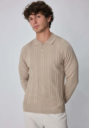 Jeune homme aux cheveux bouclés portant un polo en maille beige à manches longues et un pantalon de couleur claire, debout devant un fond uni.