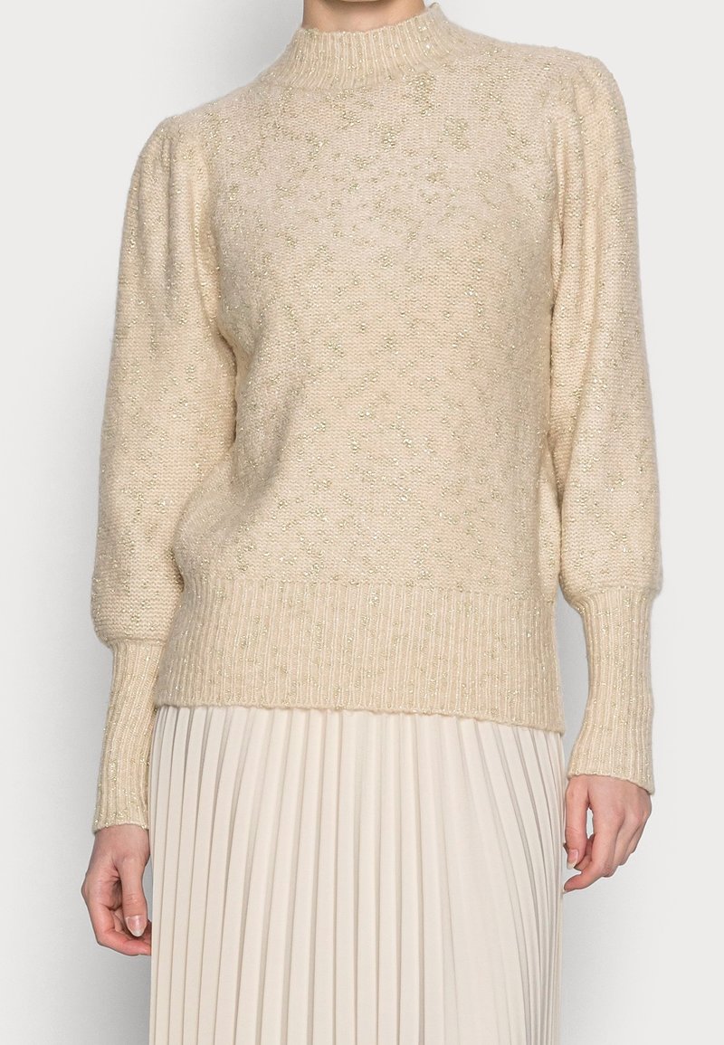 Beiger Strickpullover mit gerippten Bündchen und Saum, der eine strukturierte Oberfläche mit schimmernden Akzenten aufweist. Hoher Kragen und Puffärmel.