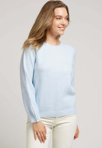 Pull en bleu clair fabriqué dans un tissu doux et texturé. Il a de longues manches et un ourlet à côtes, avec des accents de boutons au niveau des poignets.