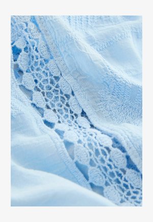 Ljust blått tyg med intrikata spetsdetaljer och texturerade mönster längs kanterna, vilket betonar mjukheten och den sköra hantverksskickligheten.