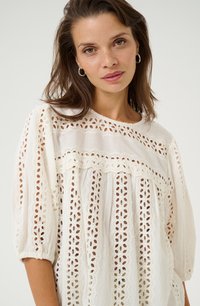 Blusa blanca con intrincados recortes de encaje, cuello redondo y mangas abullonadas. La textura suave y el diseño en capas crean una apariencia ligera y etérea.