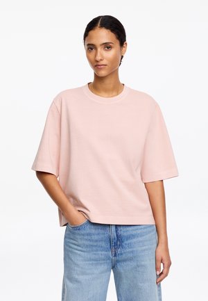 Kobieta o ciemnych włosach, ubrana w luźną, jasnoróżową koszulę z krótkim rękawem i jasnoniebieskie dżinsy, stoi z jedną ręką w kieszeni na białym tle.