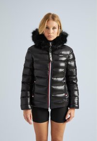 Blondýna v lesklé černé péřové bundě s kapucí z umělé kožešiny a předním zipem, oblečena do černých šortek na světlém pozadí.