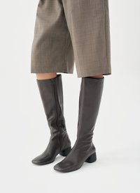 Καφέ δερμάτινες μπότες μέχρι το γόνατο με λεία υφή, τετράγωνη μύτη και χοντρό τακούνι· διαθέτουν πλευρικό φερμουάρ για εύκολη εφαρμογή.