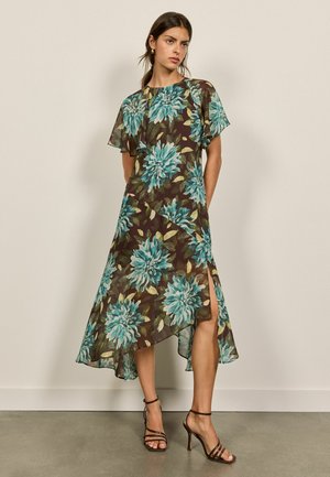 Kvinde iført en brun kjole med store blågrønne blomsterprint, flagrende ærmer, asymmetrisk hæm, sidesplit og sorte sandaler med høje hæle og remme.