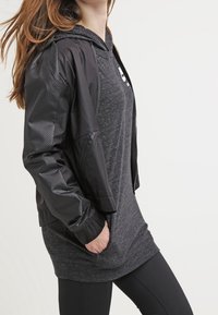 Black bomber jacket with textured sleeves over a dark gray long shirt, featuring a hood and front pockets, styled with black leggings.