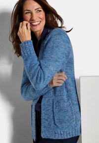 Lächelnde Frau in einer blauen Strickjacke mit Taschen, die vor einem weißen Hintergrund steht und ihr Gesicht und ihren Arm berührt.