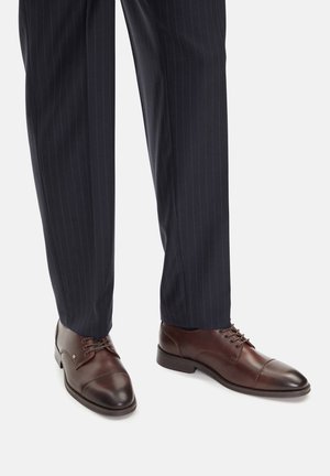 Brązowe skórzane buty wizytowe z prostokątnym noskiem, zawiązywane na sznurowadła. Połączone z ciemnymi spodniami w prążki, prezentujące formalny wygląd.