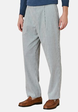 Mann trägt weiße und schwarze vertikal gestreifte Hosen mit braunen Mokassins und einem dunkelblauen Langarmhemd, steht mit entspannten Händen.