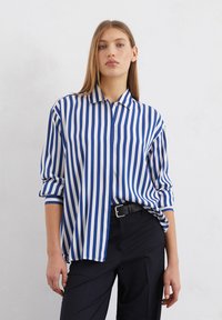 Chemise rayée bleue et blanche avec un col, un devant à boutons et des manches retroussées, fabriquée en tissu lisse, associée à un pantalon foncé.