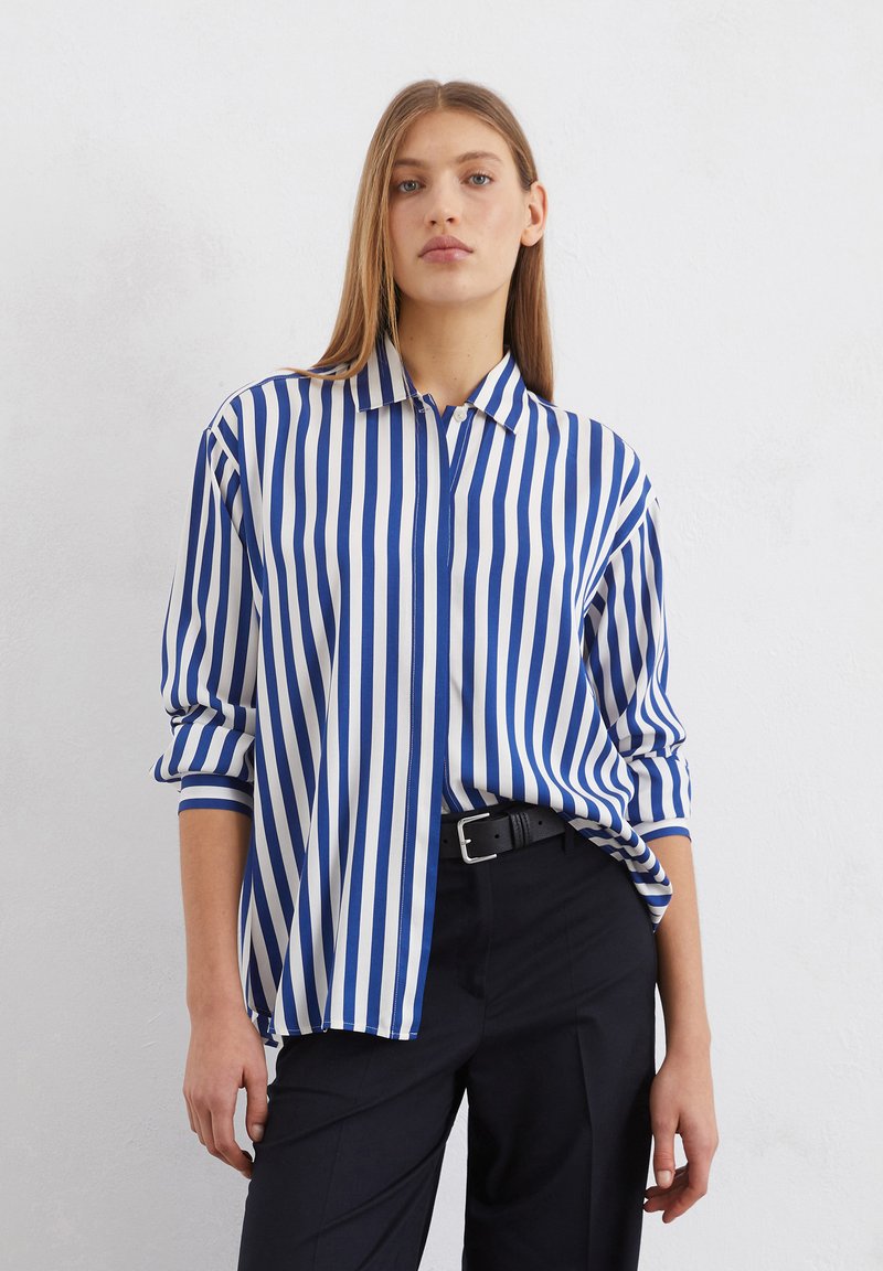 Chemise rayée bleue et blanche avec un col, un devant à boutons et des manches retroussées, fabriquée en tissu lisse, associée à un pantalon foncé.