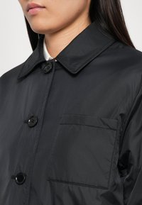 Femme portant une veste noire à boutons avec un col et une poche poitrine, avec de longs cheveux noirs visibles sur l'épaule.