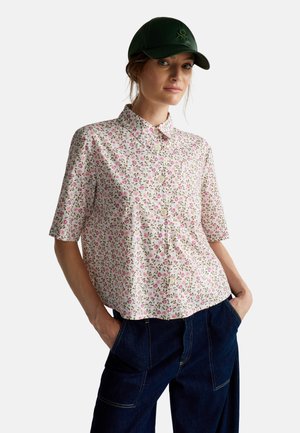 Vrouw die een bloemdessins korte mouwen overhemd draagt, donkere denim broek met handen in de zakken, en een donkergroene baseballpet.