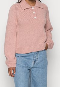 Rosa stickad tröja med en ribbad textur, krage och tre dekorativa knappar. Bärs över ljusblå denimjeans.