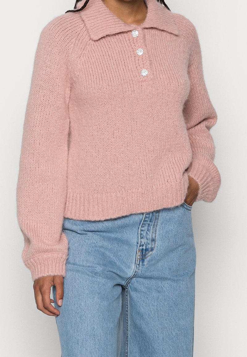 Rosa stickad tröja med en ribbad textur, krage och tre dekorativa knappar. Bärs över ljusblå denimjeans.