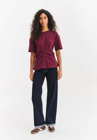 Vínové tričko s krátkým rukávem s jemným předním švem a vyšívaným detailem, doplněné širokými tmavomodrými džínami a hnědými botami.