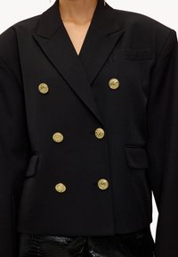 Veste blazer noire à double boutonnage avec boutons dorés, dotée d'un revers cranté et d'une poche poitrine. Le tissu semble lisse et structuré.