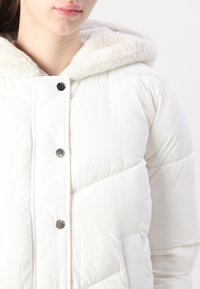 White puffer jacket with a faux fur-lined hood, snap buttons, quilted texture, and side pockets. Smooth, lightweight material.
