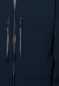 Veste bleu marine avec une texture lisse, deux poches zippées avec des accessoires noirs et une fermeture éclair centrale sur toute la longueur.