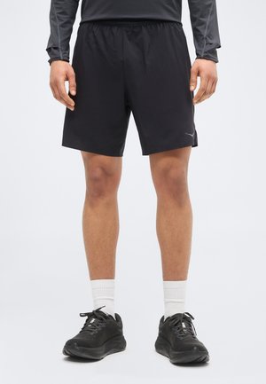 Man draagt zwarte sportshorts, zwarte hardloopschoenen met witte sokken, en een donkergrijs shirt met lange mouwen, staand tegen een effen achtergrond.