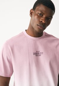 Heleroosa lühikeste varrukatega puuvillane T-särk, millel on keskel rinnal tumesinises kirjas tekst "SHIBUYA CITY TOKYO JAPAN".