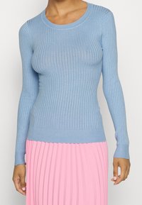 Světlomodrý žebrovaný svetr s dlouhými rukávy a přiléhavým střihem, doplněný plisovanou růžovou sukní. Hladká textura, kulatý výstřih.