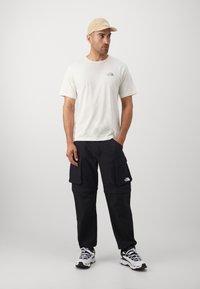 White short-sleeve t-shirt with a green logo, black cargo pants with pockets, and beige cap. Shoes are black and white athletic style.