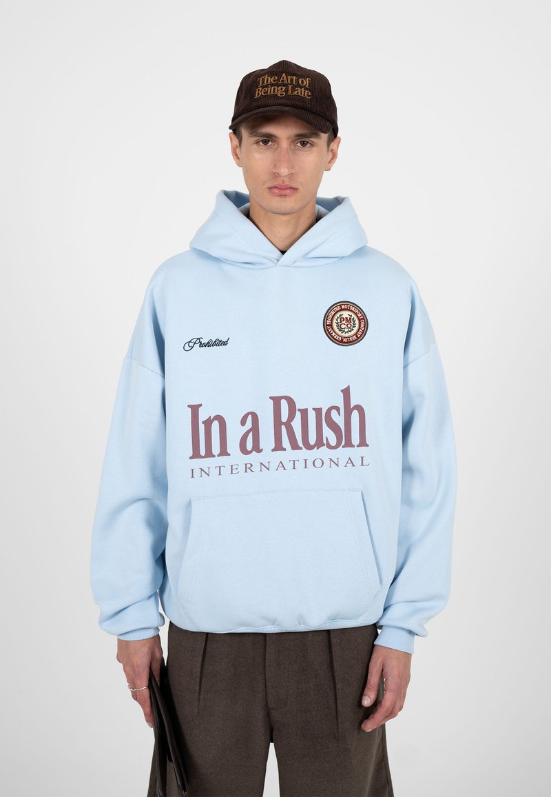 Homme portant un sweat à capuche bleu clair avec le texte « In a Rush International », une casquette marron sur laquelle est inscrit « The Art of Being Late » et un pantalon foncé.