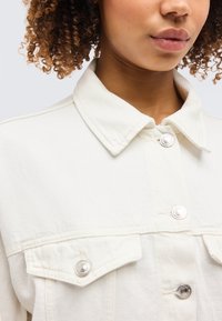 Νεαρό άτομο με σγουρά μαλλιά που φοράει λευκό τζιν μπουφάν με ασημένια κουμπιά, ο γιακάς είναι ορατός και το φόντο είναι ουδέτερο.