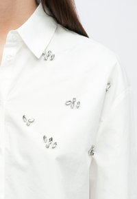 Weißes Hemd mit klassischem Kragen, verziert mit silbernen Schmucksteinen in verschiedenen Formen und Größen, die vorne aufgenäht sind.