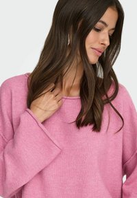 Różowy, oversize'owy sweter o fakturowanym splocie, z szerokimi mankietami i okrągłym dekoltem. Materiał wydaje się miękki i przytulny.