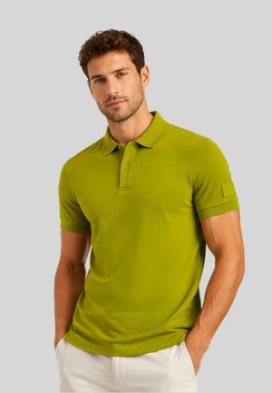 Uomo che indossa una polo verde a maniche corte con colletto e bottoni, in piedi con le mani nelle tasche di pantaloni bianchi contro uno sfondo semplice.