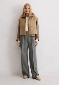 Beige gelaagde jas met twee frontzakken, gecombineerd met lichtgrijze wijde broek. Model draagt lichtbeige top en beige sneakers.