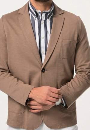 Mand iført beige blazer over vertikalt stribet skjorte, justerer sølvfarvet armbåndsur på venstre håndled, mod en ensartet lys baggrund.