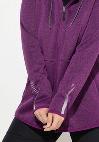 Lila Fleecejacke mit Reißverschluss vorne, Kapuze und reflektierenden Akzenten an den Ärmeln. Dicke Textur und Seitentaschen für Funktionalität.