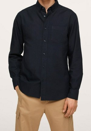 Uomo in camicia nera a maniche lunghe con taschino e pantaloni cargo beige, in piedi con una mano in tasca.