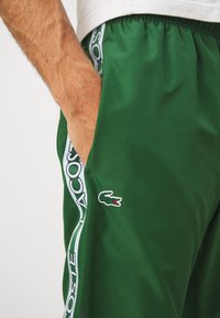 Pantaloni sportivi verdi realizzati in tessuto testurizzato, con una banda laterale bianca recante la scritta "LACOSTE" e un piccolo patch logo sul davanti.