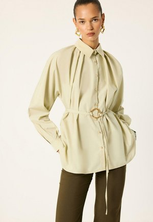 Femme portant un chemisier beige boutonné avec une ceinture cintrée à la taille et un pantalon marron ajusté, accessoirisée de grandes boucles d’oreilles géométriques dorées.