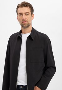 Zwarte jas met ritssluiting en kraag, met een borstzak en een gladde textuur, gedragen over een wit shirt. Eenvoudig, modern ontwerp.