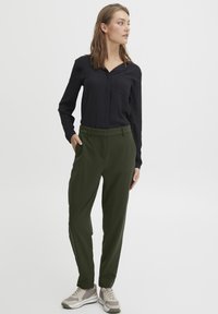 Svart langermet skjorte med krage og lomme, kombinert med olivengrønne bukser. Modellen bruker grå joggesko for et uformelt utseende.