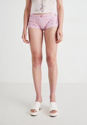 Woman wearing light pink sequined shorts, white platform slide sandals, and a sheer white top with floral details, standing on a plain white floor.