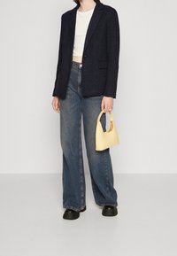 Námořnický texturovaný blazer přes bílý crop top, doplněný světle modrými širokými džínami a malou žlutou kabelkou. Černé robustní boty dokončují vzhled.