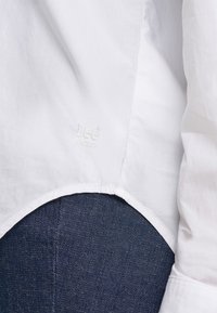 Λευκή ραφή πουκάμισου με διακριτικό κεντημένο λογότυπο Lee πάνω από σκούρο μπλε τζιν ύφασμα.