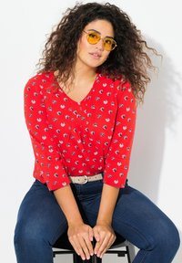 Rote Bluse mit floralen Mustern, dreißigsten Ärmeln, V-Ausschnitt und kleinen Blumenmotiven, kombiniert mit dunkelblauen Jeans und einem beigen Gürtel.