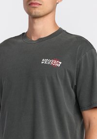 T-shirt en coton noir gris avec un petit logo Volcom blanc et rouge sur la poitrine gauche, un col crew et des manches courtes.