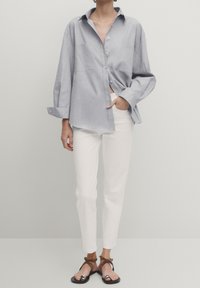 Chemise boutonnée bleu clair à coupe décontractée et manches longues, associée à un pantalon blanc ajusté et des sandales noires avec des accents marron.