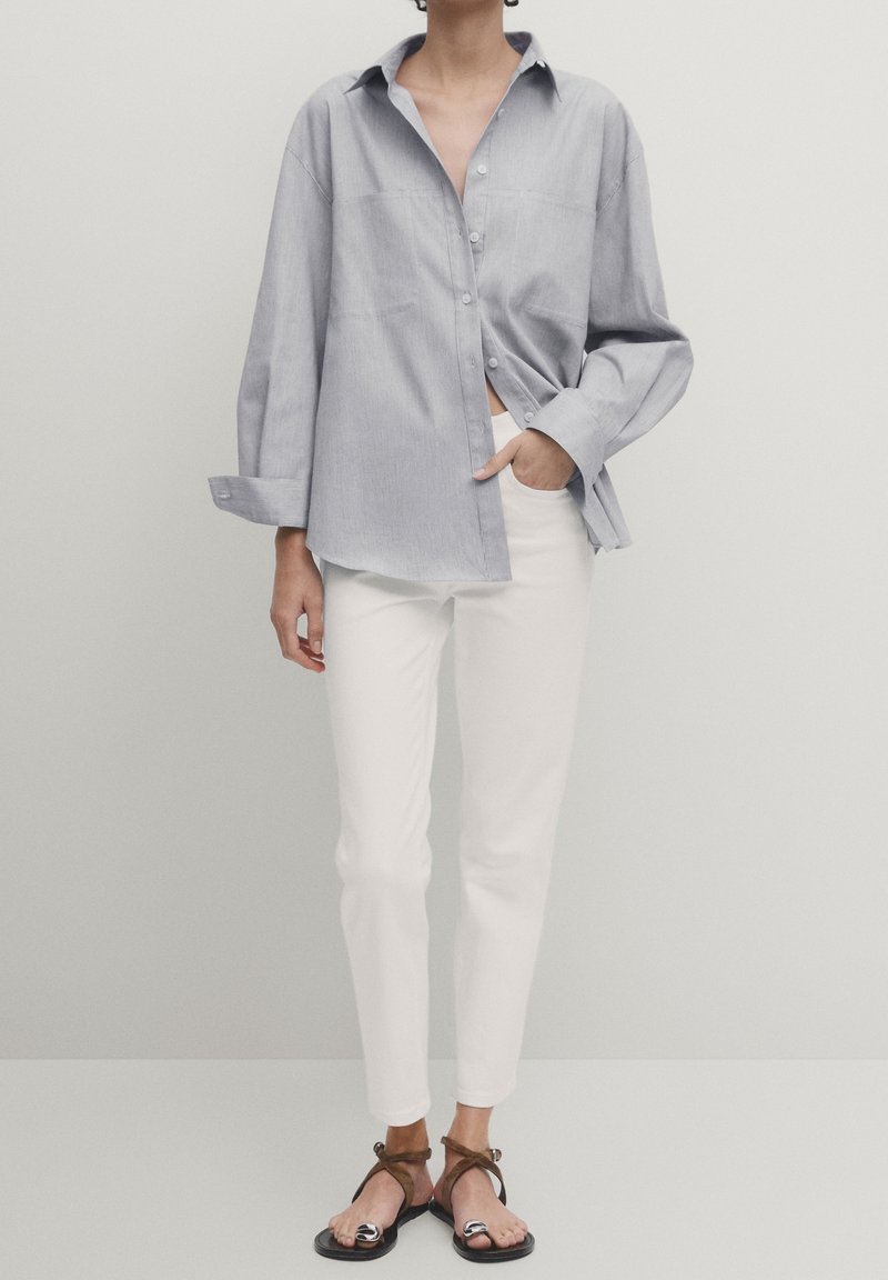 Chemise boutonnée bleu clair à coupe décontractée et manches longues, associée à un pantalon blanc ajusté et des sandales noires avec des accents marron.