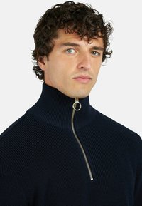 Maglione a maglia blu scuro con colletto alto e zip dorata. Tessuto strutturato e design a coste. Presenta un tirante circolare per la zip.
