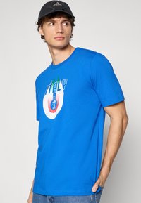 Jauns vīrietis, kas valkā melnu Adidas cepuri un zilu T-kreklu ar Itālijas grafiku, skatās uz kreiso pusi, viena roka ir kabatā.