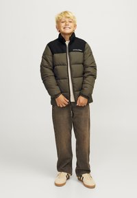 Doudoune olive avec un col noir et un logo, associée à un haut beige clair et un pantalon large marron, ainsi qu'à des baskets beige.